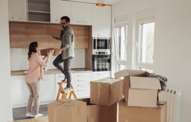 Genç çift, karton kutuları mutfaktaki yüksek elementlerde depoluyor, konsept olarak taşınıyor ve eşyaları açıyor.