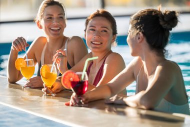 Üç kadın havuzda dinleniyor, gülüyor ve sıcak havanın tadını çıkarırken renkli kokteyller yudumluyorlar. Enerjik atmosfer onların neşeli ruhlarını güçlendirir.