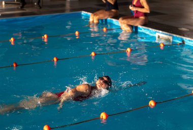 Yüzücüler kapalı havuzda idman yapıyorlar, eğitim sırasında suda süzülürken gösteri becerileri sergiliyorlar. Parlak ışıklandırma onların hareketlerini vurgular.