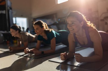 Bir grup güzel kadın kapalı spor salonunda hasırın üzerinde kalas yapıyor.