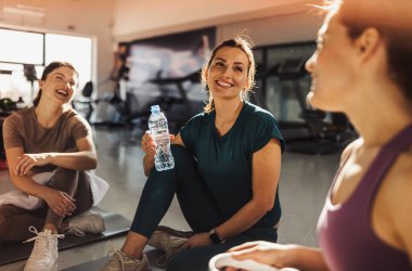 Spor salonunda oturmuş spor yaptıktan sonra gülüp sohbet eden üç kadın. Biri su şişesi tutuyor, bir anlık dostluk ve zindelik gösteriyor.