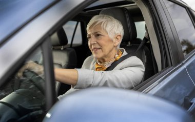 Üst düzey bir kadın açık havada araba sürerken gülümser, direksiyonun başındaki deneyimin güvenini ve zevkini gösterir.