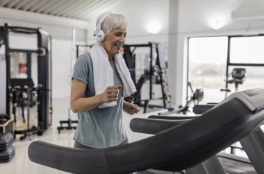 İyi donanımlı spor salonunda koşu bandında koşan, kulaklık takan ve boynuna havlu takmış, spor yapmaya odaklanmış yaşlı bir kadın..