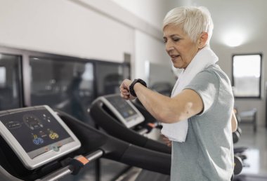 İyi donanımlı spor salonunda koşu bandında koşan, kulaklık takan ve boynuna havlu sarılmış yaşlı bir kadın akıllı saate bakıyor..
