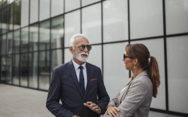 Takım elbiseli ve güneş gözlüklü kıdemli bir adam modern binanın dışında iş elbiseli genç bir kadınla sohbet ediyor ve profesyonel bir diyalog sergiliyor..