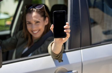 Neşeli kadın sürücü, güneşli bir günde arabasında otururken akıllı telefonunu kaldırır. Anın tadını çıkarır ve diğerleriyle iletişim kurar.