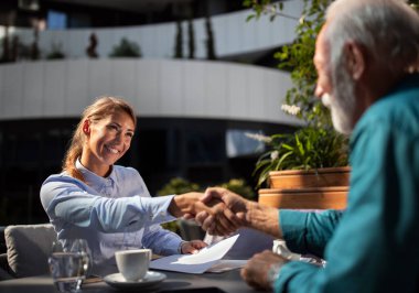 Genç bir kadın açık bir kafede yaşlı bir adamla el sıkışıyor, ikisi de profesyonel giyinmiş. Kahve içerken iş görüşmesi yapıyor gibiler..