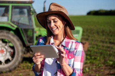 Kadın işçi, güneşli bir günde açık mavi gökyüzünün altında arka planda traktörle yeşil alanda tablet kullanırken gülümsüyor..