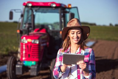 Kadın işçi, güneşli bir günde açık mavi gökyüzünün altında arka planda traktörle yeşil alanda tablet kullanırken gülümsüyor.