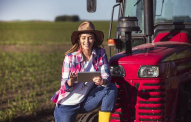 Kadın işçi, güneşli bir günde açık mavi gökyüzünün altında traktöre yaslanarak yeşil alanda tablet kullanırken gülümsüyor..