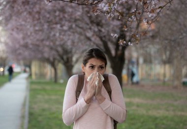 Genç güzel bir kadın hapşırıyor ve şehir parkında çiçek açan ağacın önünde burnunu siliyor. Bahar poleni alerjisi krizi.