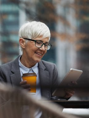 Yaşlı iş kadını dijital tabletle ilgili belgeleri inceliyor ve kafenin dışında otururken bir şeyler içiyor.
