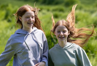 Uzun kızıl saçlı gülümseyen iki kız gülüyor ve parlak güneş ışığı altında yeşil alanda koşuyor, neşe ve dostluk anlarını yakalıyorlar.