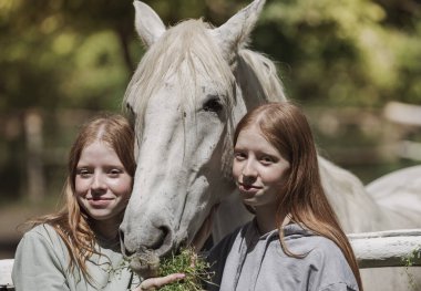 İkiz kardeşler gülümser ve görkemli beyaz atlarla görkemli açık havada çarpışırlar, yemyeşil ve sıcak güneş ışığıyla çevrili.