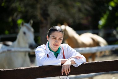 Kadın veteriner portresi, güneşli bir ahırda ahşap çitin yanında duruyor. Arka planda iki atı gözlemliyor ve hayvan sağlığına olan bağlılığını gösteriyor.