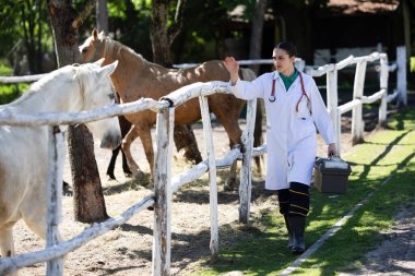 Beyaz önlüklü ve çizmeli veteriner otlakta atlara yaklaşır, yeşilliklerle çevriliyken sağlık çantası taşır.