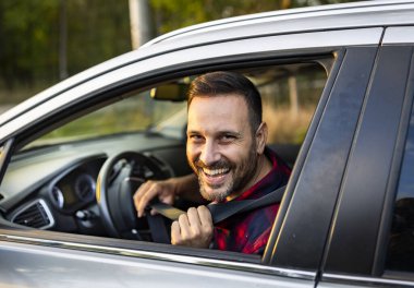 Adam park halindeki arabada otururken emniyet kemerini bağlarken parlak bir şekilde gülümsüyor. Sıcak güneş hoş bir açık havada parıldıyor..