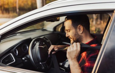 Adam park halindeki arabada otururken emniyet kemerini bağlarken parlak bir şekilde gülümsüyor. Sıcak güneş hoş bir açık havada parıldıyor..