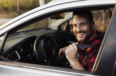 Adam park halindeki arabada otururken emniyet kemerini bağlarken parlak bir şekilde gülümsüyor. Sıcak güneş hoş bir açık havada parıldıyor..