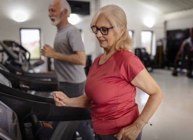 Kardiyo yapan kendinden emin yaşlı bir kadın yanında eşiyle koşu bandı kullanıyor.