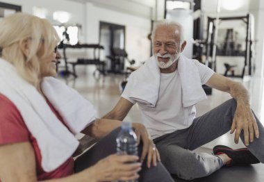 Spor salonundaki yoğun egzersizden sonra yerde oturan yaşlı çift ellerinde su şişesi ve omuzlarında havlular.