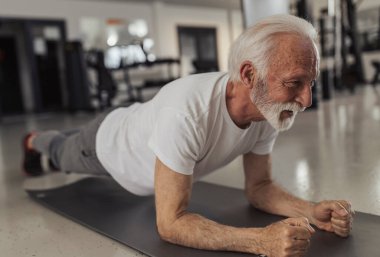 Spor salonunda yaşlı bir adam, tahta döşeme yapıyor, yerde dayanıklılık eğitimi alıyor.