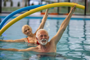 Yaşlı adam ve kadın kafalarının üstünde havuz eriştesi tutuyorlar ve kapalı havuzda su aerobiği yapıyorlar.
