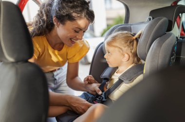 Kadın, güneşli bir günde arabanın arka koltuğunda otururken genç kızına emniyet kemeri takmasında yardım ediyor. Güvenlik ve bakım çok açık.