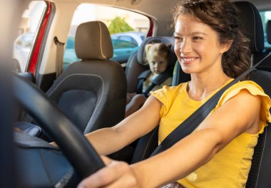 Gülümseyen anne arabasını güvenle sürerken, küçük çocuğu güneşli bir günde arka koltukta güvenli bir şekilde oturuyor.