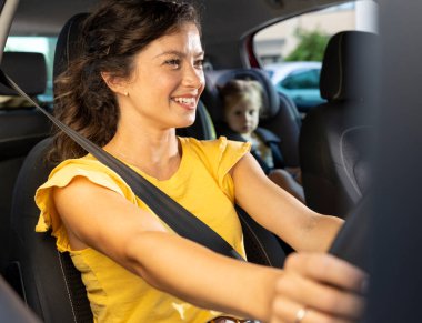 Gülümseyen anne arabasını güvenle sürerken, küçük çocuğu güneşli bir günde arka koltukta güvenli bir şekilde oturuyor.