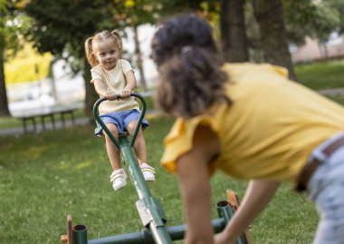 Genç kız, annesi ağaçlarla çevrili, güneşli bir ortamda dengede dururken parkta tahterevalli ile oynamaktan hoşlanıyor.