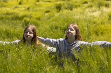 Tek yumurta ikizi kızlar bahar güneşinin tadını çıkarırken uzun çimenlerde oturuyorlar, elleri açık ve gözleri kapalı.