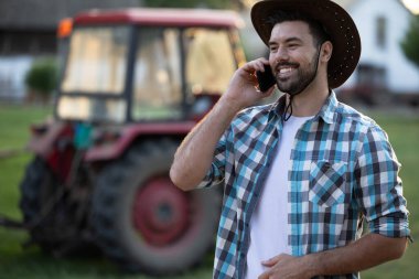 Güneşli tarlada akıllı telefonunda kareli gömlek ve şapka konuşmaları giyen çiftçi, traktör arka planda kırsal alanda park halinde.