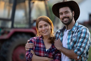 Neşeli çift çiftlikte birbirine yakın duruyor, arkada traktör var. Kadın ve erkek kameraya bakıyor.