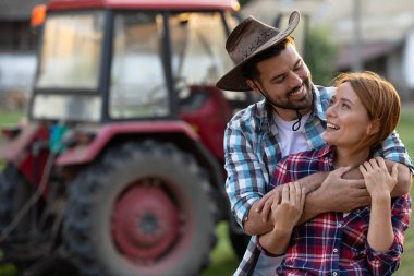 Neşeli çift çiftlikte birbirine yakın duruyor, arkada traktör var. Adam kovboy şapkası takıyor ve kız arkadaşına bakıyor.