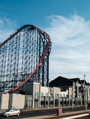 A towering roller coaster looms over urban architecture under a clear blue sky. clipart