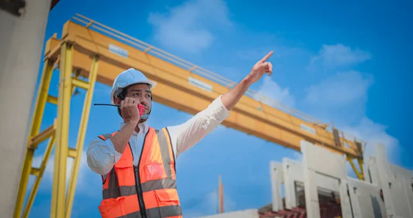 Professional Asian male civil engineer or architect wear safety helmet. While working using walkie-talkie talking to someone checking list project on construction site. Real estate concept.