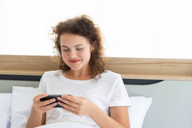 Genç, güzel, mutlu bir kadın sabah uyandıktan sonra yatakta uzanırken rahatlıyor ve akıllı telefon kullanıyor..