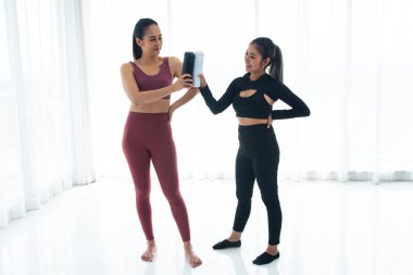 Siyah spor giyimli, kendine güvenen bir kadın, parlak, minimalist bir arka plana karşı bordo giysili arkadaşına su şişesini sunuyor.