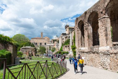 Roma, İtalya-17 Nisan 2024: Roma 'daki Roma Forumu' nu gezen turistler, antik harabeler ve tarihi yapılar arasında yürüyor. Sahne eski ve yeni karışımı yakalar, yüksek tuğla kemerler, taş sütunlar, ve yemyeşil parçalı bulutlu altında