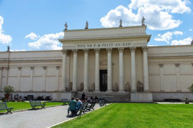 Vatikan, İtalya-18 Nisan 2024: Vatikan Müzeleri 'nin büyük binasının önünde oturan turistler, klasik sütunlar ve heykeller yer alıyor..