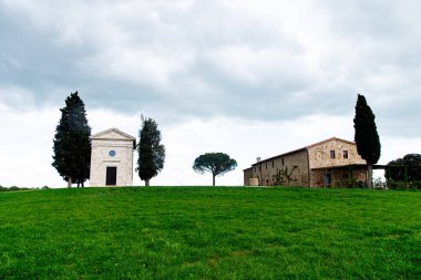 Toskana, İtalya-22 Nisan 2024: Tuscany, İtalya 'da bulutlu bir gökyüzünün altında çimenli bir tepede bulunan sevimli küçük bir şapel ve taştan ev.