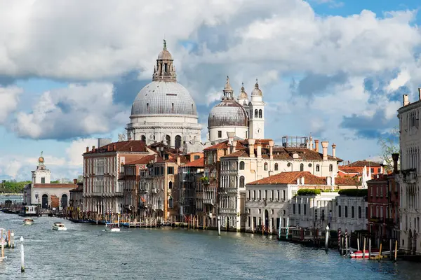 stock image Venice, Italy-April 24, 2024: A picturesque view of the Basilica di Santa Maria della Salute with historic buildings lining Venice's Grand Canal under a bright, cloudy sky.