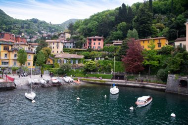 Como Gölü, İtalya-26 Nisan 2024: Renkli evleri, yemyeşil alanları ve canlı bir gökyüzünün altında kıyıya yanaşmış tekneleri olan büyüleyici göl kenarı köyü..