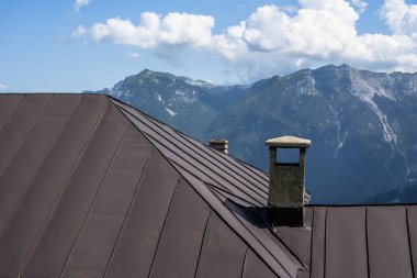 Güneşli bir günde şafak vakti, arka planda dağ tepeleri ve baca kazanı olan kahverengi yapraklı, metal bir dağ sığınağı çatısı.