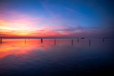 Venedik gölünün sakin sularına kırmızı ve turuncu renklerle yanan günbatımı yansıtılır. Venedik 'teki Venedik Lido' sundan çekilen tuğla ve ağaçlarla.
