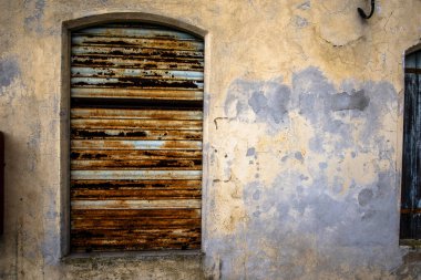 Çürüyen alçı duvarda eski paslı metal pencere perdesi, soyulmuş boya ve yıpranmış doku. Patina, yaş ve kırsal karakterle dolu cesur bir mimari detay..