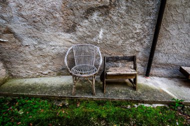 İki kırsal sandalye hasır ve bir ahşap yan yana, yosunlu beton bir yüzey üzerindeki eski dokulu bir duvara karşı, klasik ve kırsal atmosferi olan sessiz, nostaljik bir açık hava manzarası.