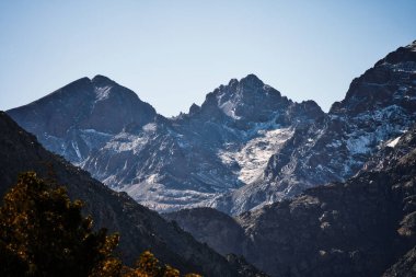 Fas 'taki görkemli Atlas Dağları. Açık mavi gökyüzü altında karla kaplı tepeler ve engebeli arazi nefes kesici, dramatik bir manzara yaratır..