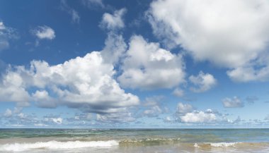 Sahil manzarası sakin deniz ve mavi gökyüzü ile beyaz bulutlar, sakin ve rahatlatıcı bir sahne yaratıyor.